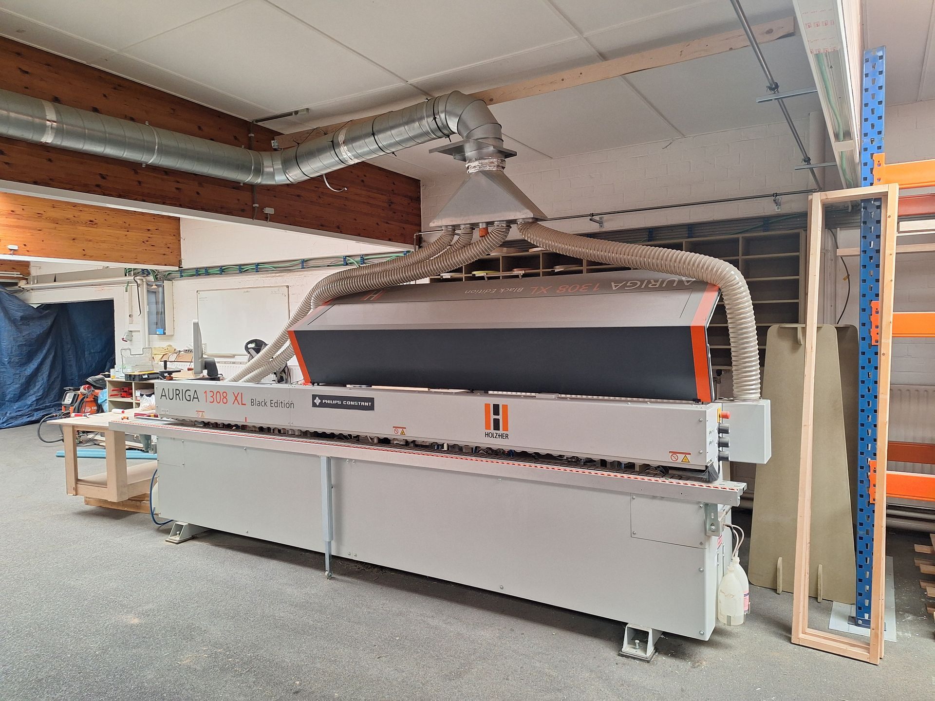 Een grote industriële houtbewerkingsmachine in een werkplaats, met ventilatiebuizen. De kleuren grijs en oranje zijn prominent aanwezig.