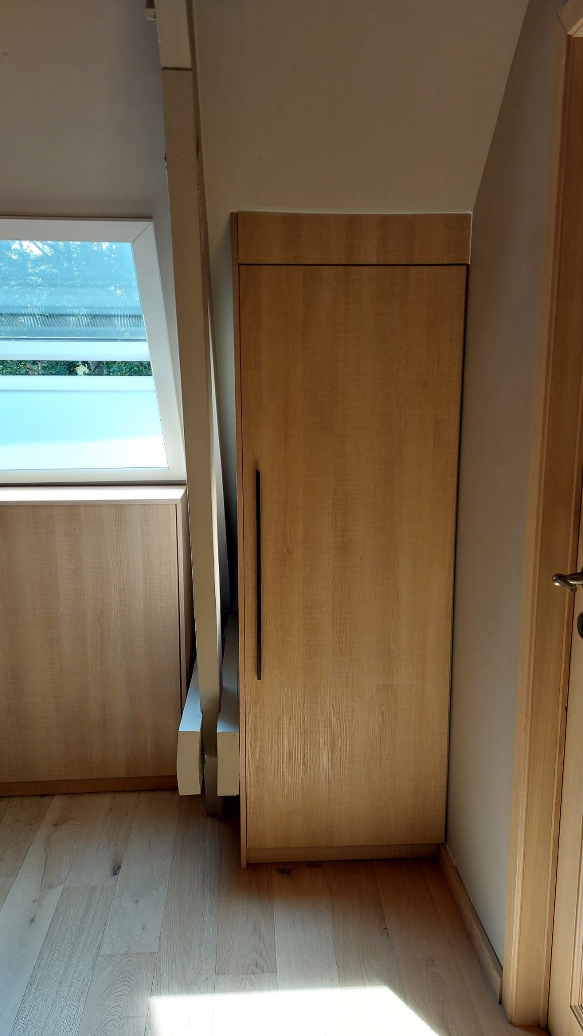 Een kast van licht hout ingebouwd in een schuine muur, naast een raam en een deur.