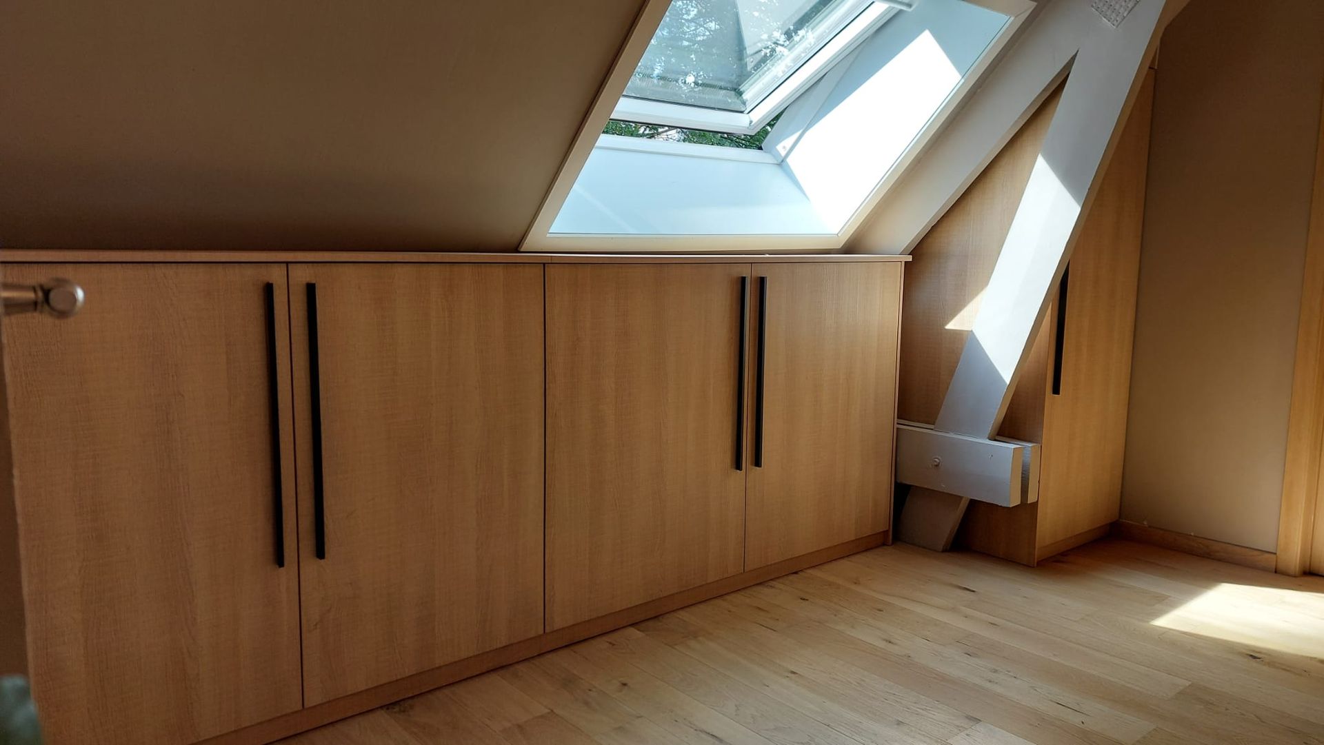 Ingebouwde houten kasten met zwarte handgrepen onder een schuin plafond en een dakraam in een kamer met houten vloer.