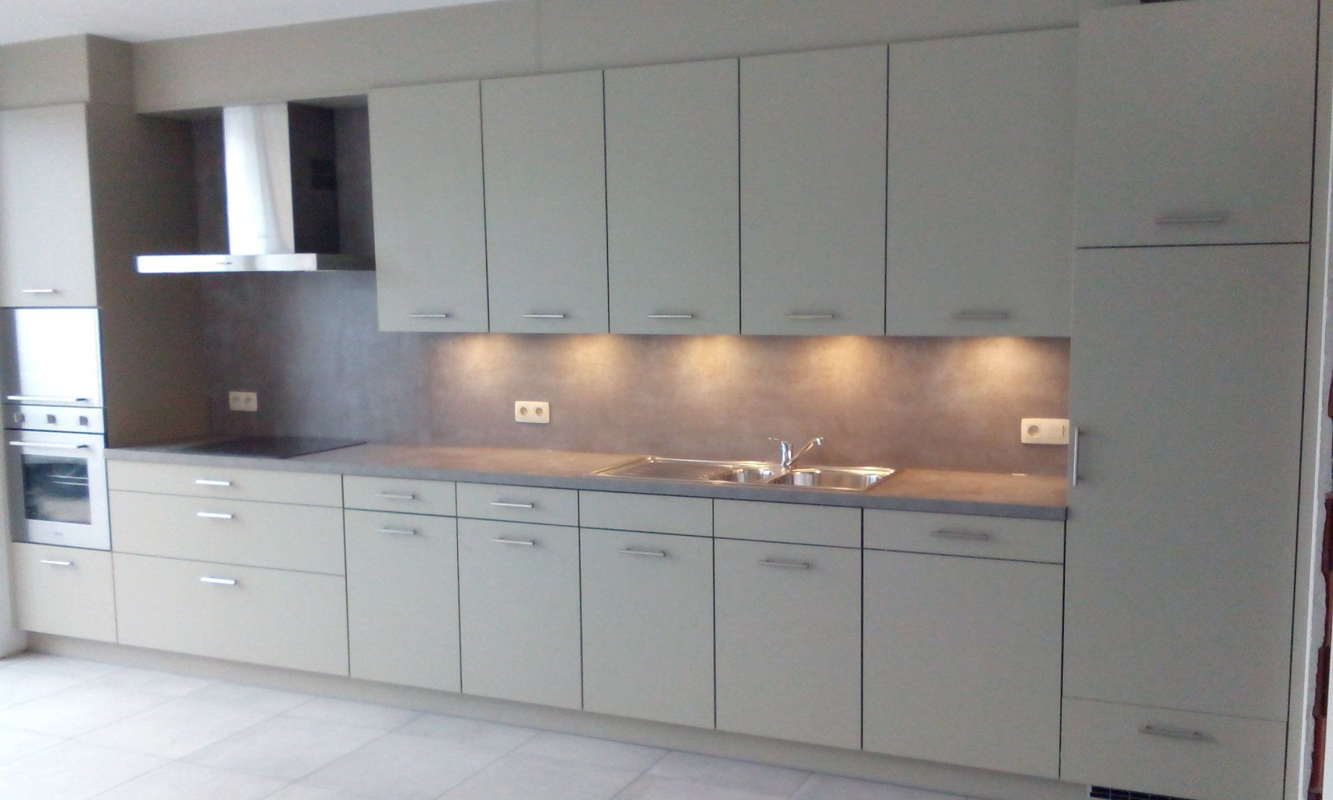 Beige keuken met bovenkastjes, aanrechtblad en inbouwapparatuur. Grijze spatwand en verlichting onder de kastjes.