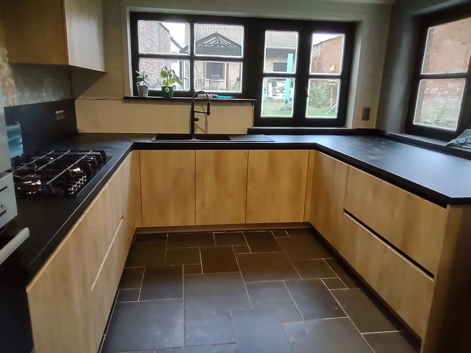 U-vormige keuken met lichte houten kasten, zwarte aanrechtbladen en een raam met donkere kozijnen. Donkere tegelvloer.