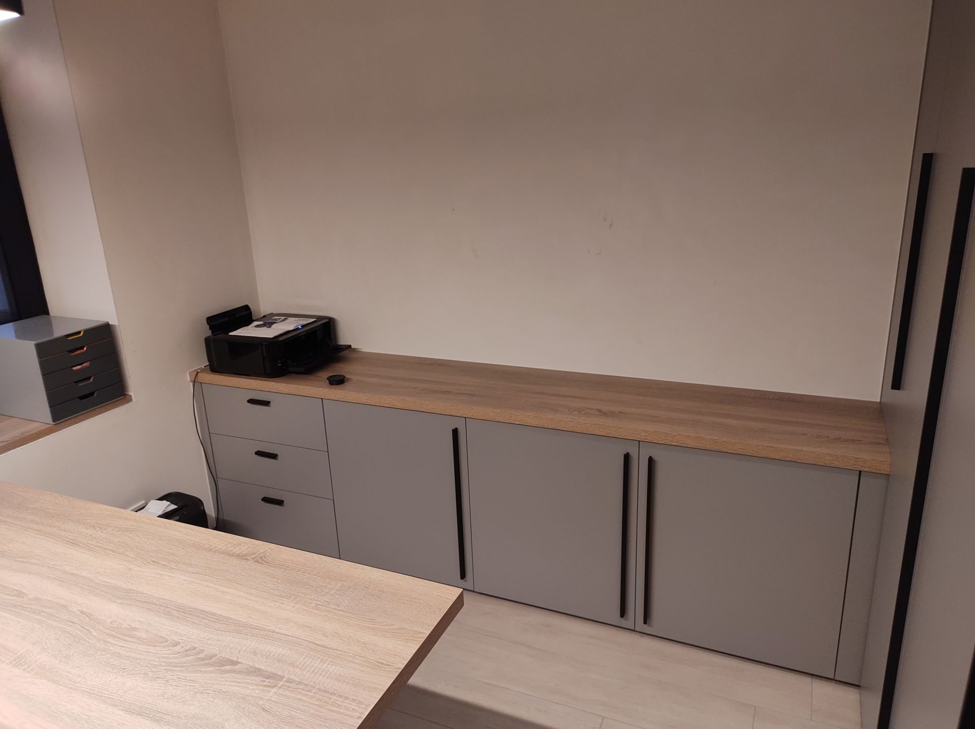 Ingebouwde grijze opbergkast met lades en deuren, met een lichthouten aanrechtblad. Op het aanrecht staat een printer.