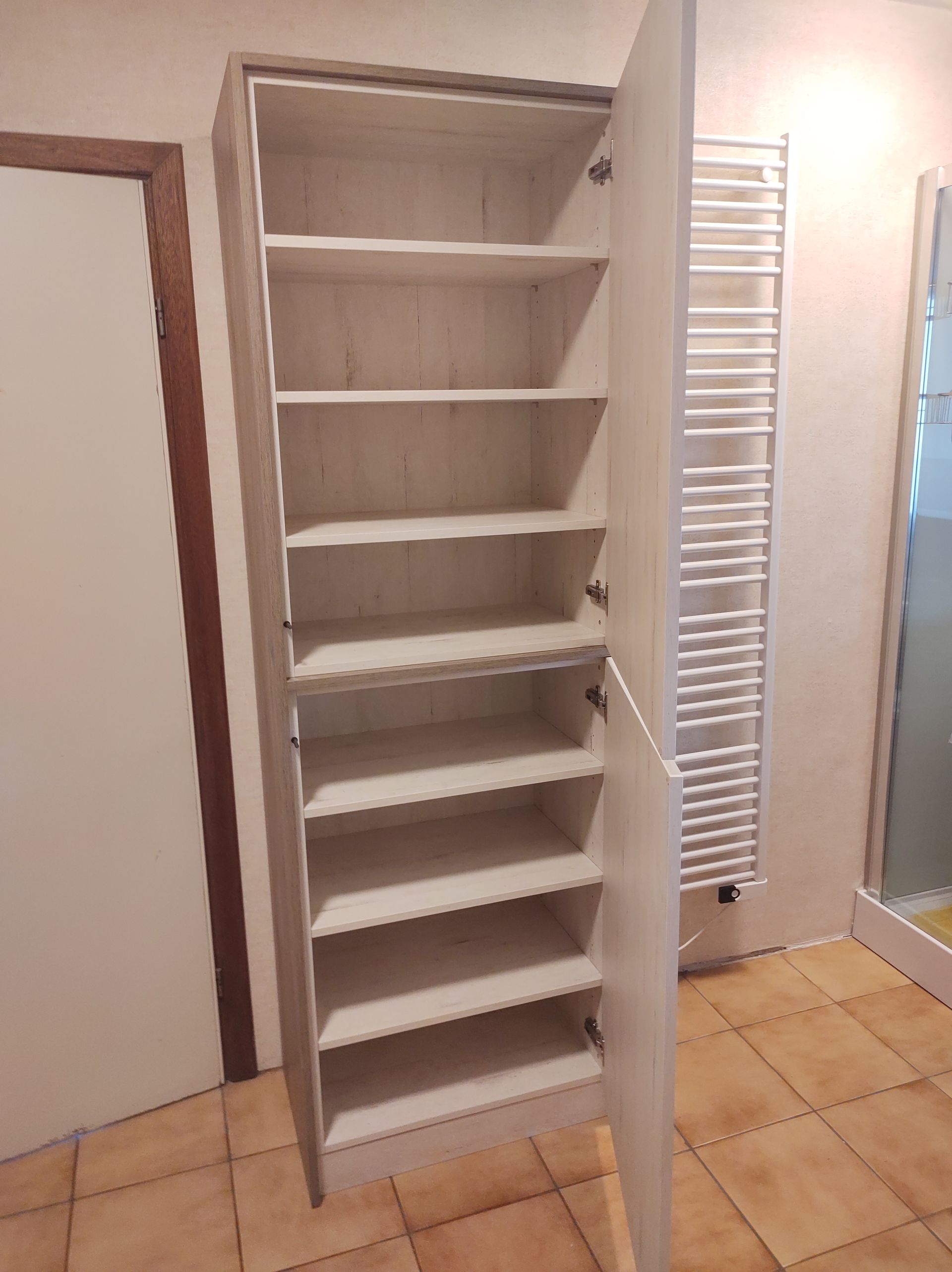 Hoge, witte kast met een open deur waaruit meerdere planken tevoorschijn komen, in een kamer met een tegelvloer en een handdoekwarmer.