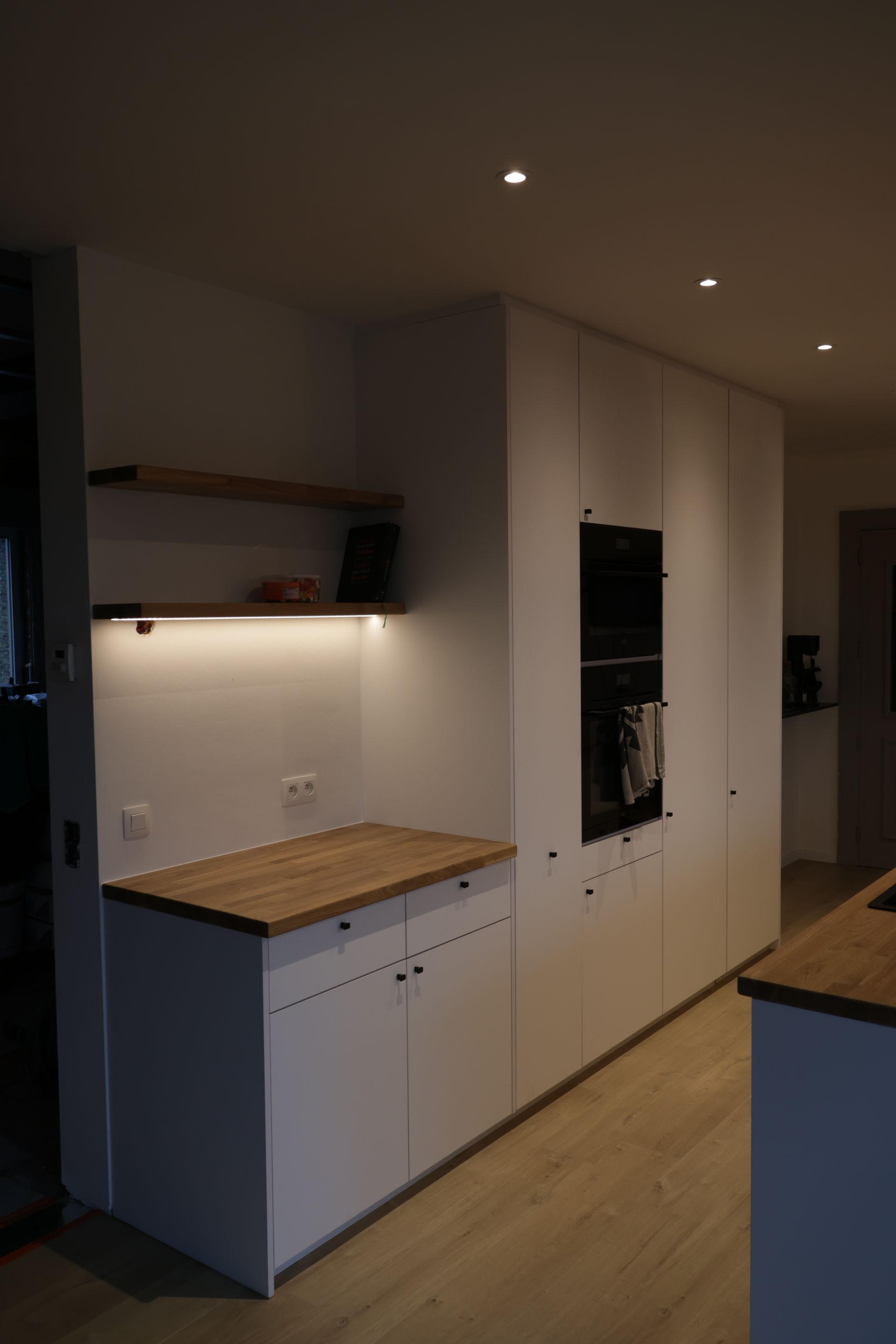 Moderne witte keuken met houten aanrechtbladen en planken. Ingebouwde oven en minimalistisch design.