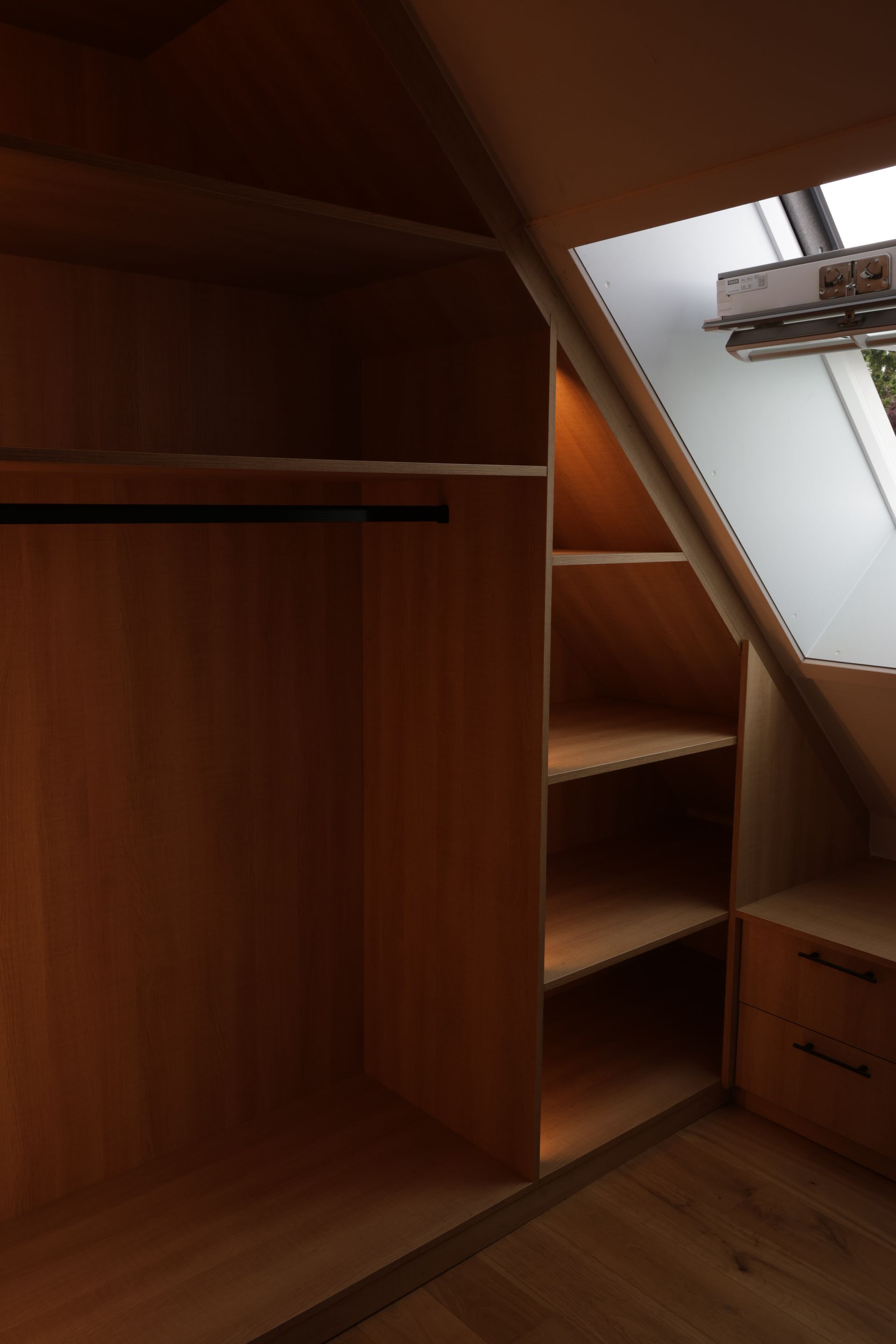 Houten kast ingebouwd in een schuin plafond. De kast heeft planken, een hangroede en lades. Natuurlijk licht stroomt door een raam.