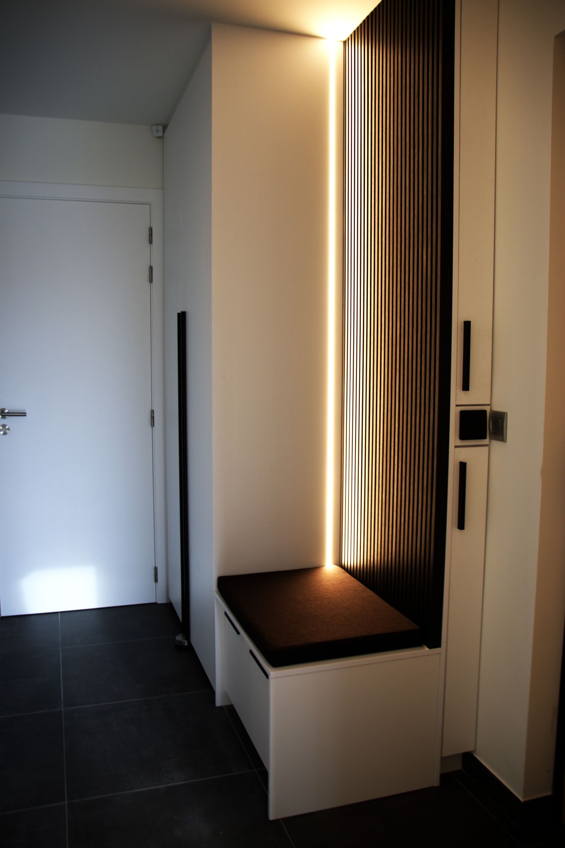 Een moderne entree met een ingebouwde bank, verticale houten lambrisering en verborgen verlichting. Links is een witte deur.