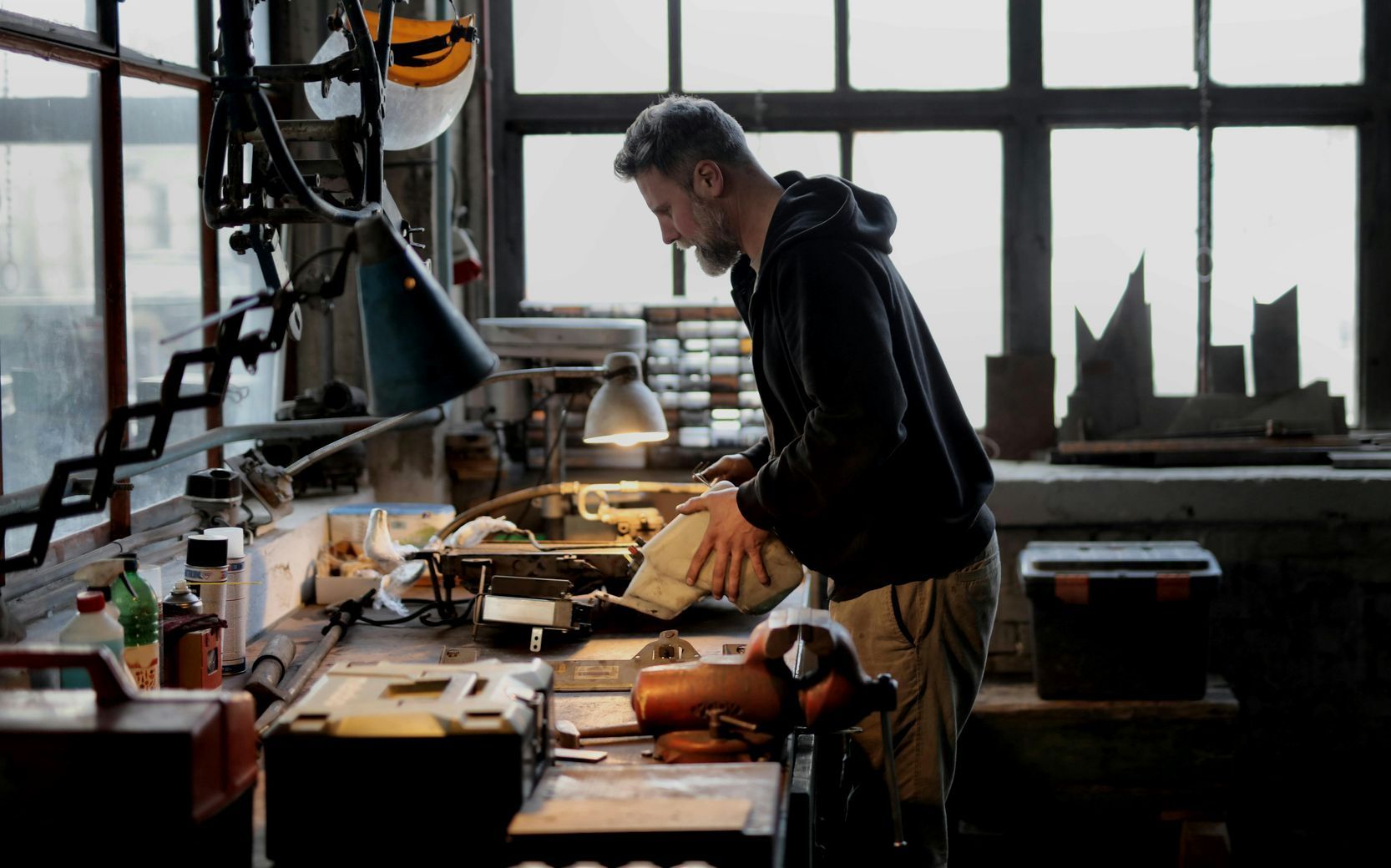 Een man werkt aan een werkbank in een werkplaats, verlicht door een lamp. Hij onderzoekt een voorwerp, naast een bankschroef.