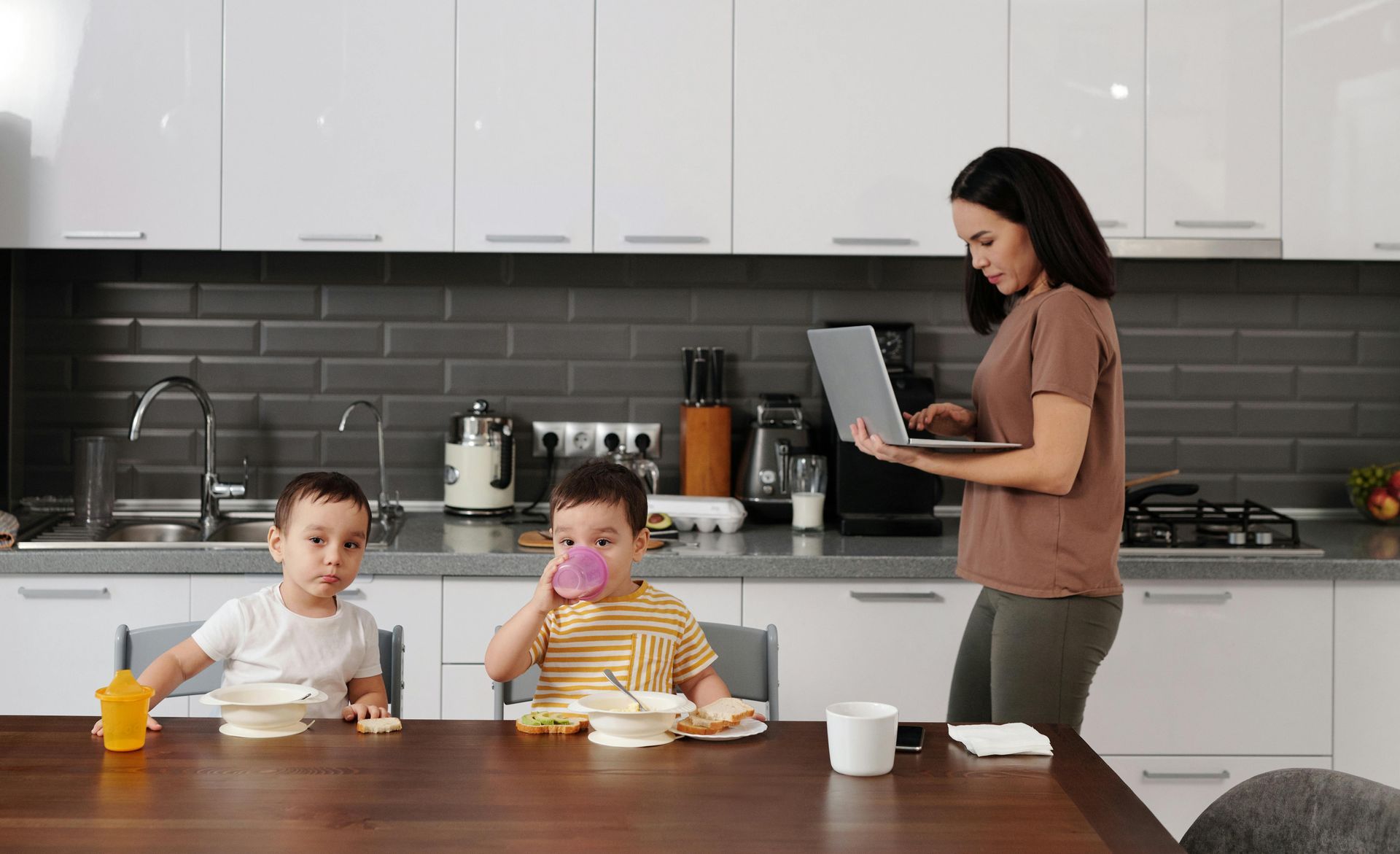 Een vrouw werkt op een laptop in de keuken, terwijl twee jonge kinderen aan tafel ontbijten.