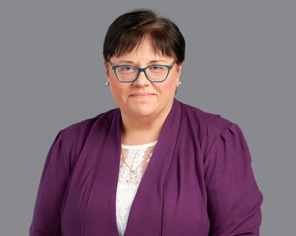 A woman wearing glasses and a purple jacket is standing in front of a gray background.