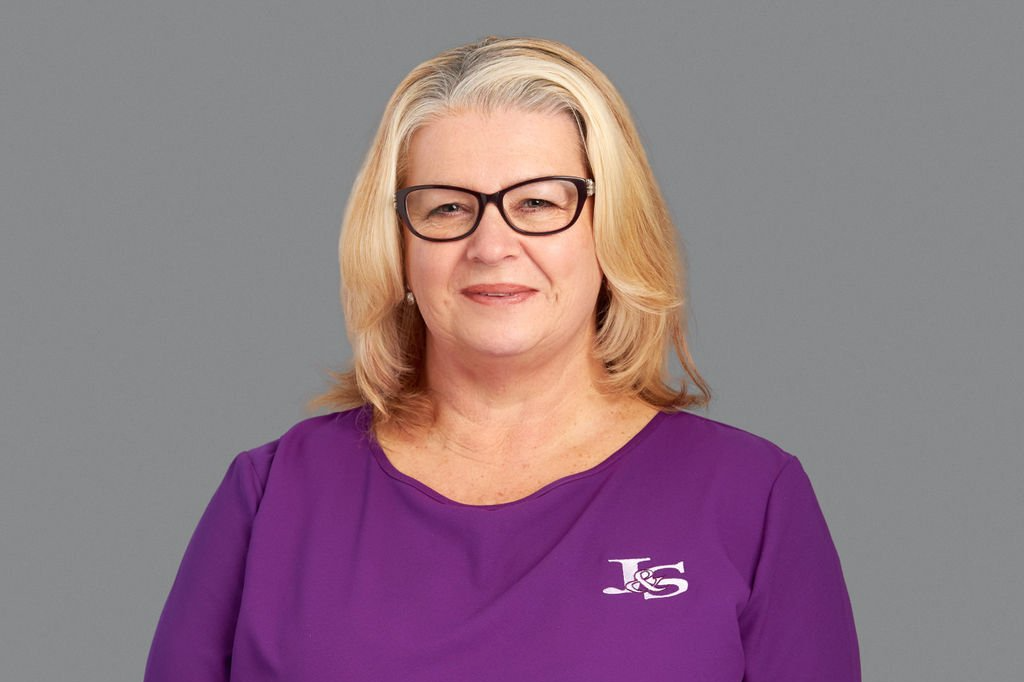 A woman wearing glasses and a purple shirt is standing in front of a gray background.