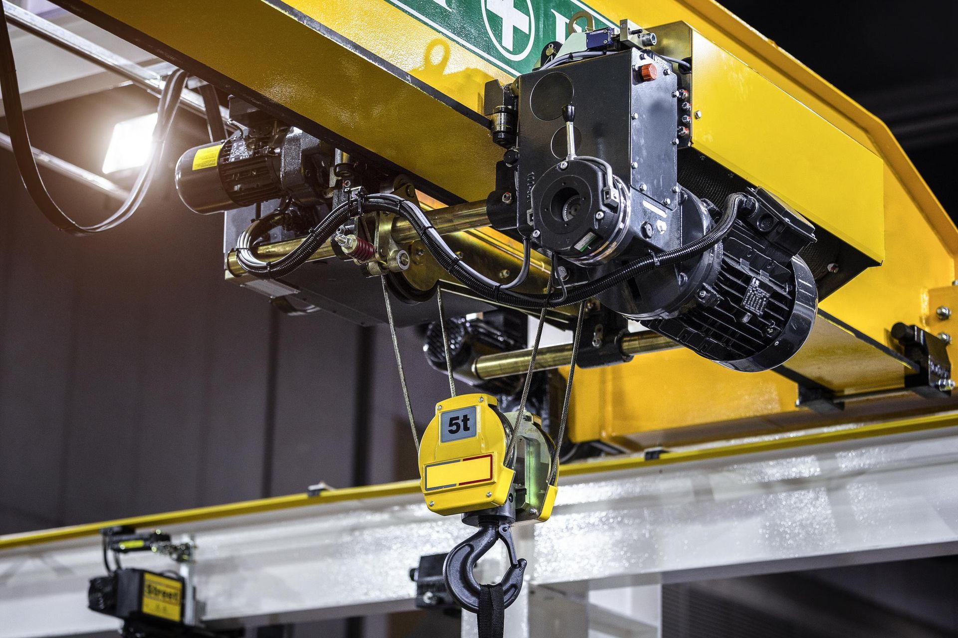 Yellow overhead crane in a factory, with a black hoist and hook.