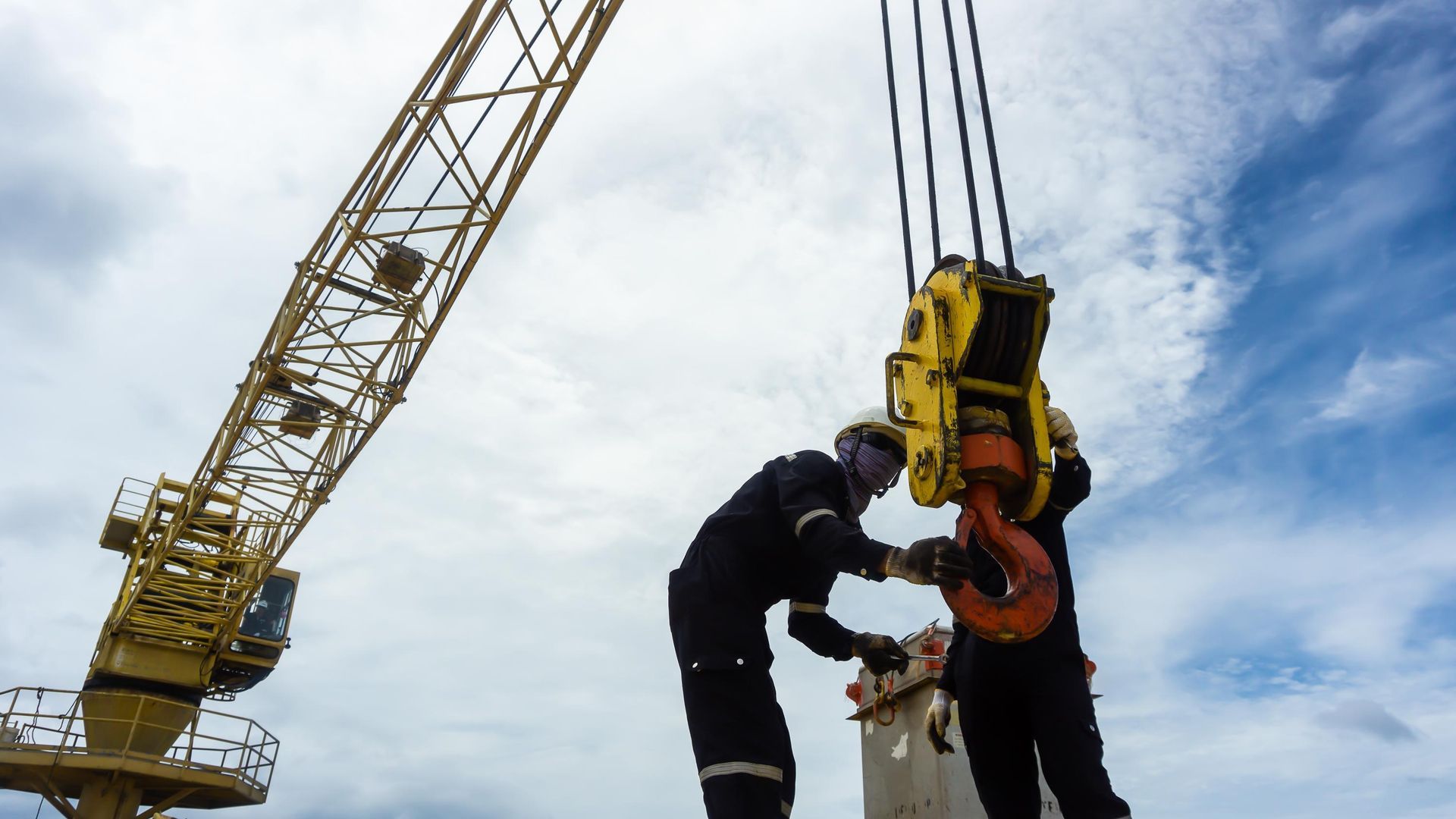 Workers connecting load to crane hook outdoors, cloudy sky overhead.