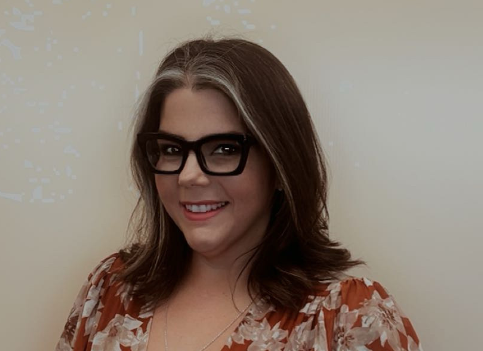 Woman with glasses, smiling. Brown hair with grey streaks, floral top. Against a beige wall.