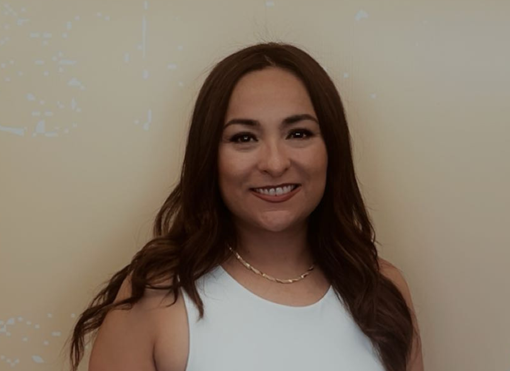 Woman with brown hair smiling, wearing a white top and gold necklace, against a beige wall.