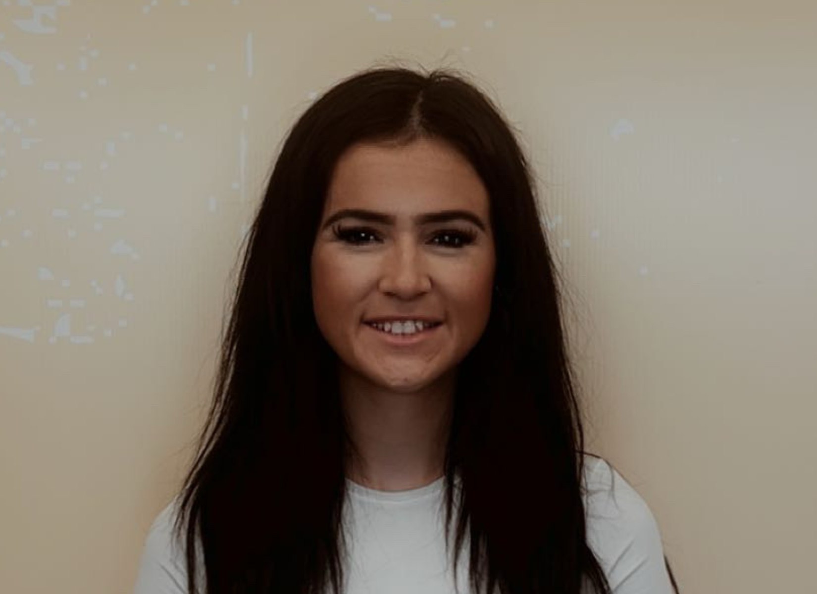 Woman with dark hair smiling, wearing white shirt, in front of a beige wall.