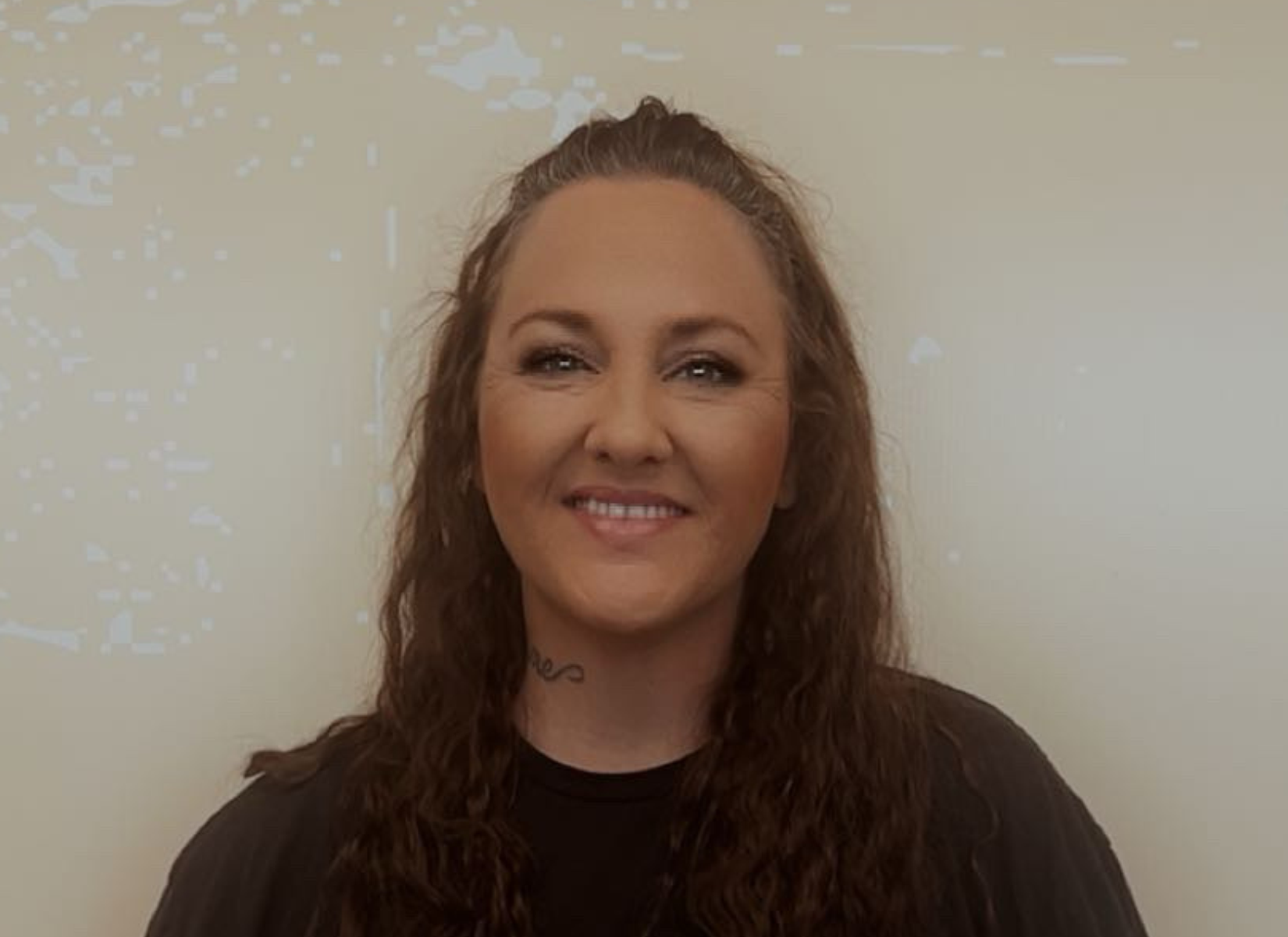 Woman with long, wavy brown hair smiles; wearing black top, standing in front of a plain wall.