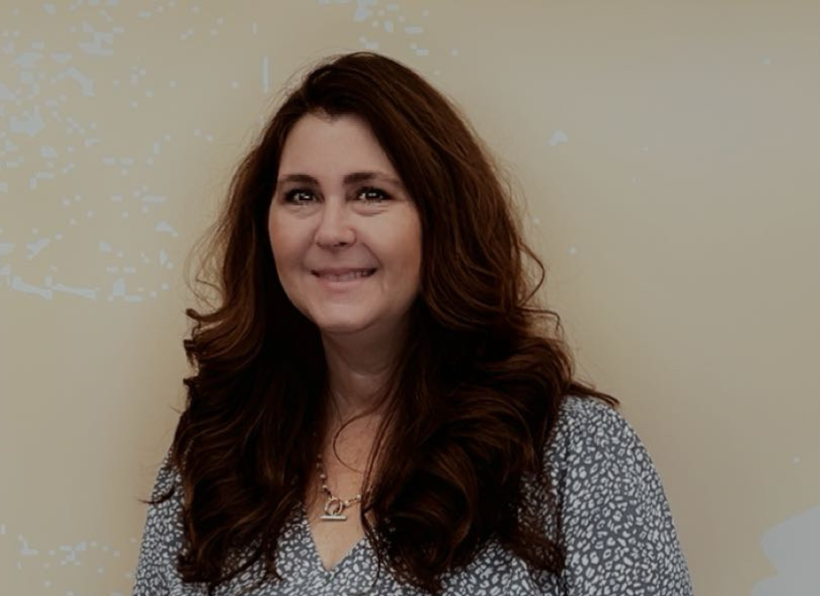 Woman with long brown hair smiles, wearing a patterned top, stands against a beige wall.