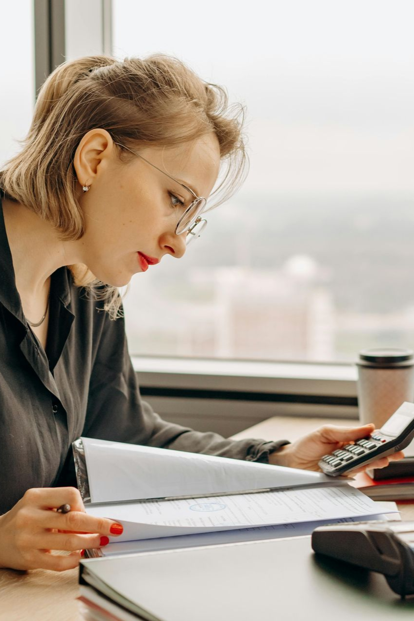 Woman in glasses working at a desk, using a calculator and reviewing documents.