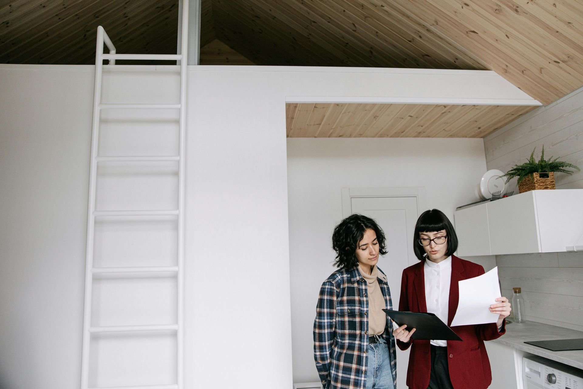 Two people look at documents in a minimalist room with a ladder, white walls, and wood ceiling.