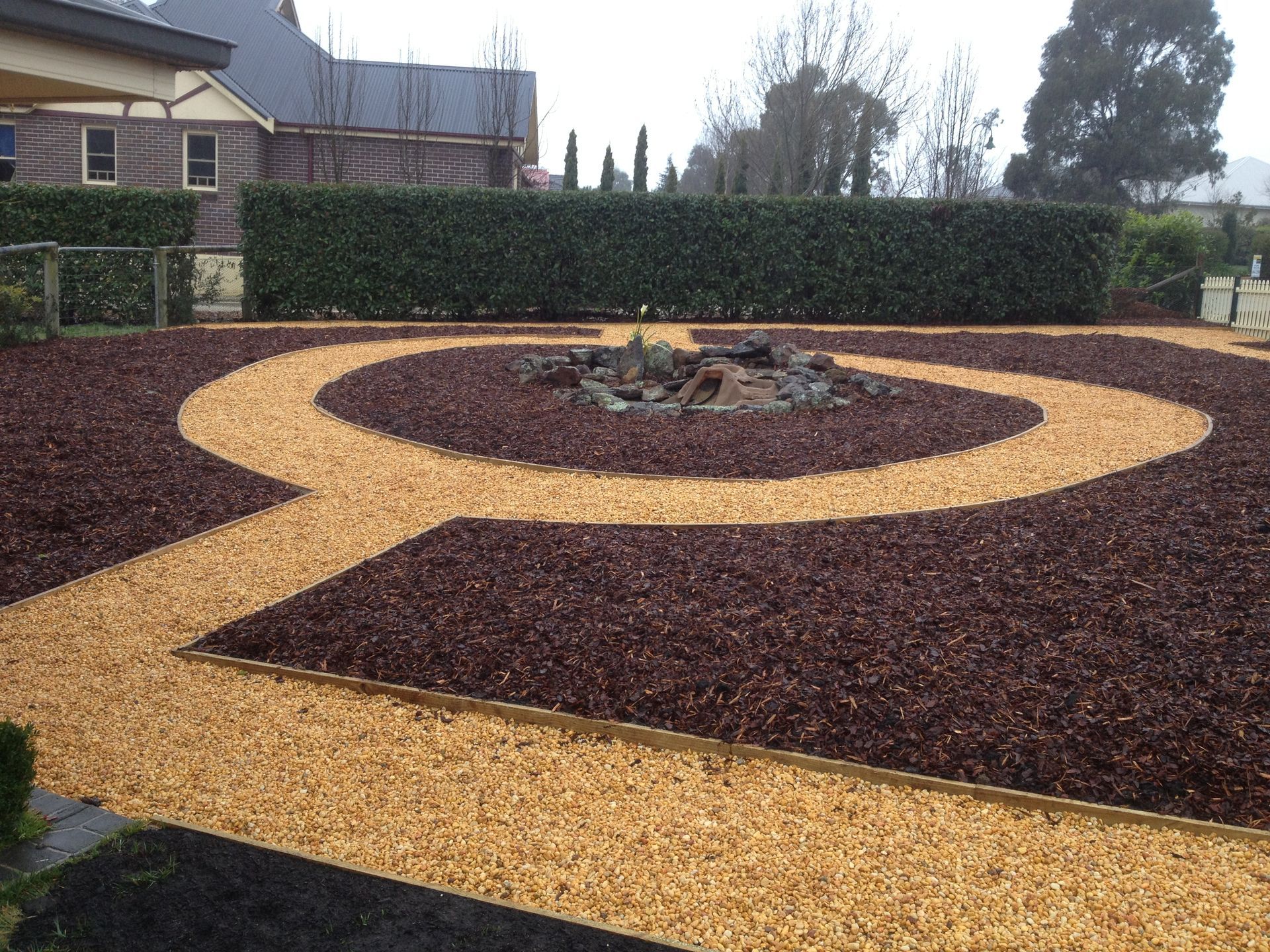A person in green overalls prunes a bush with a yellow handled tool. Grass and landscaping in view.