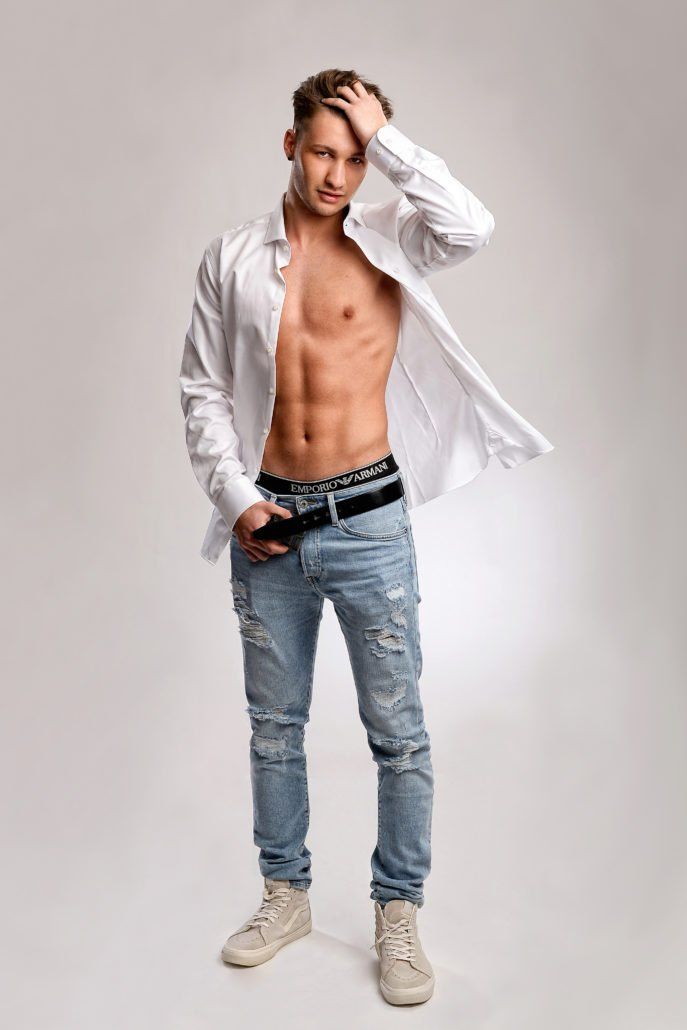 Ein Mann ohne Hemd, in einem weißen Hemd und blauen Jeans steht vor einem weißen Hintergrund.