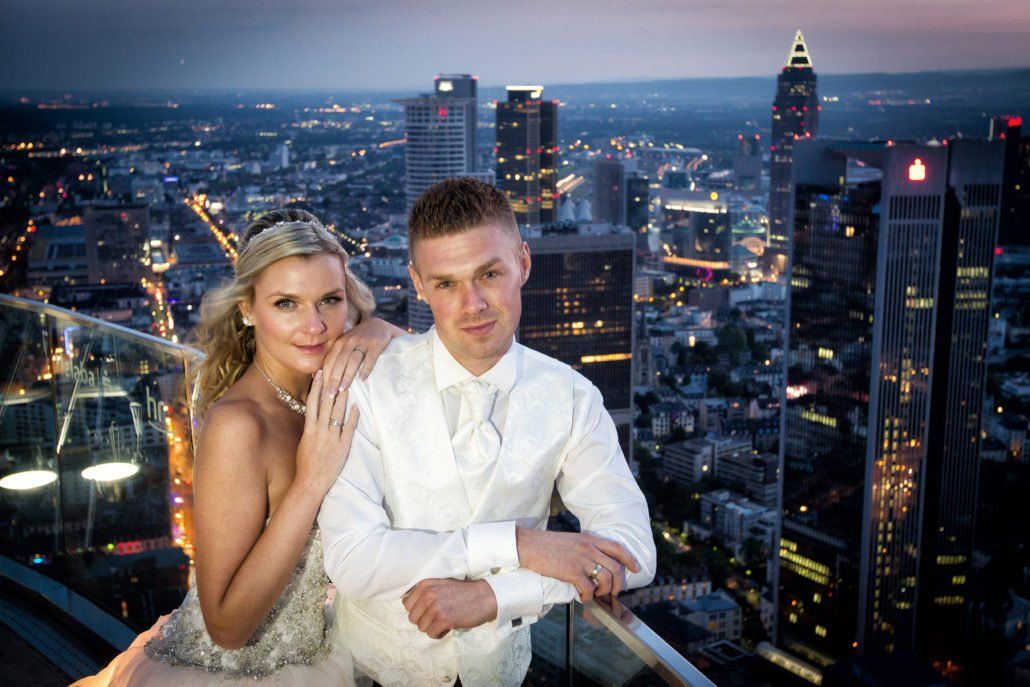 Eine Braut und ein Bräutigam posieren für ein Foto auf einem Balkon mit Blick auf eine Stadt bei Nacht.