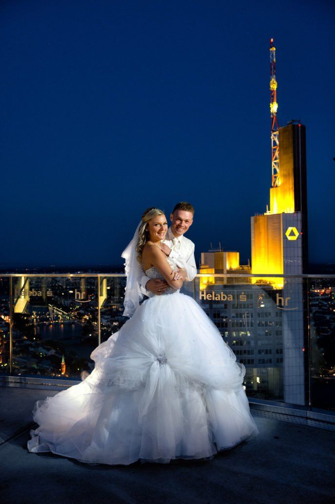 Eine Braut und ein Bräutigam posieren für ein Foto auf einem Balkon mit einem hohen Gebäude im Hintergrund.