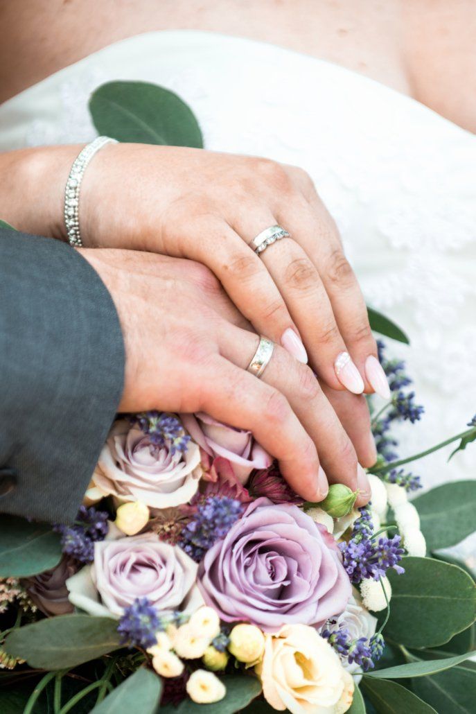 Eine Braut und ein Bräutigam halten über einem Strauß lila Blumen die Hände.