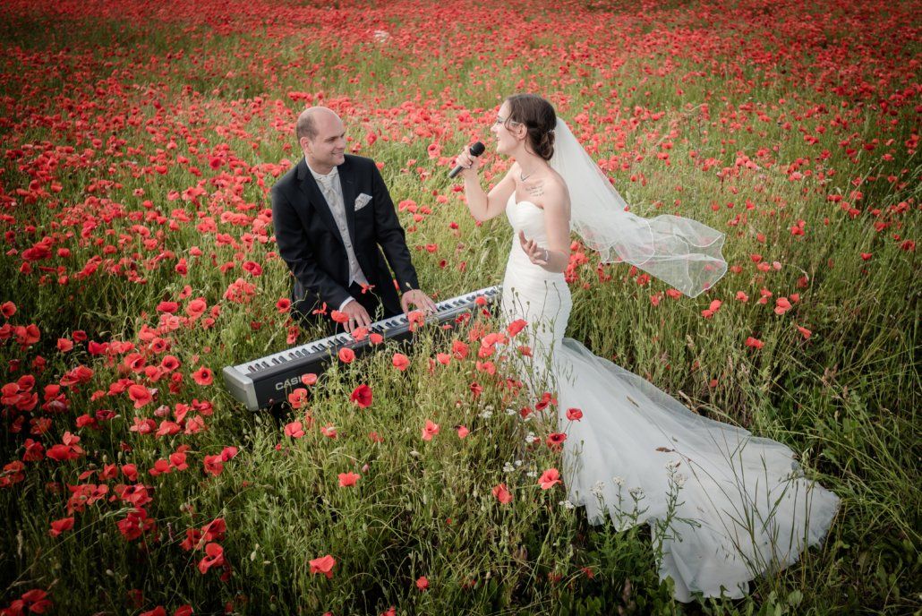 Eine Braut singt in ein Mikrofon, während ein Bräutigam auf einem Feld voller roter Blumen auf einem Keyboard spielt.