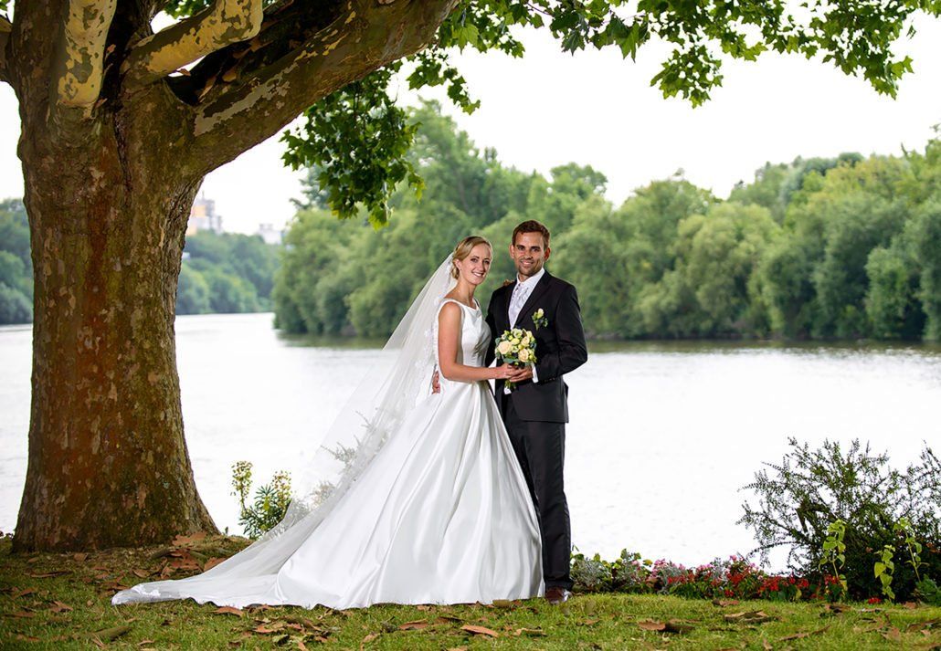 Eine Braut und ein Bräutigam posieren für ein Foto unter einem Baum in der Nähe eines Sees.