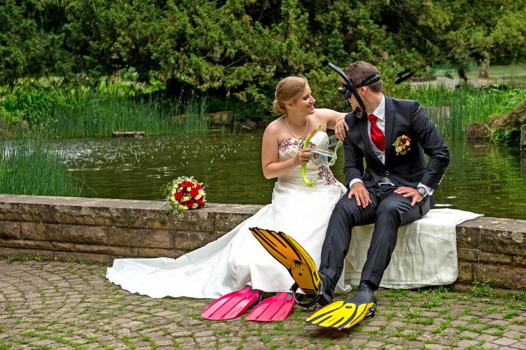 Eine Braut und ein Bräutigam sitzen auf einer Backsteinmauer neben einem Teich.