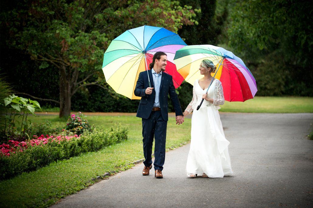 Eine Braut und ein Bräutigam gehen mit regenbogenfarbenen Regenschirmen in den Händen einen Weg entlang.