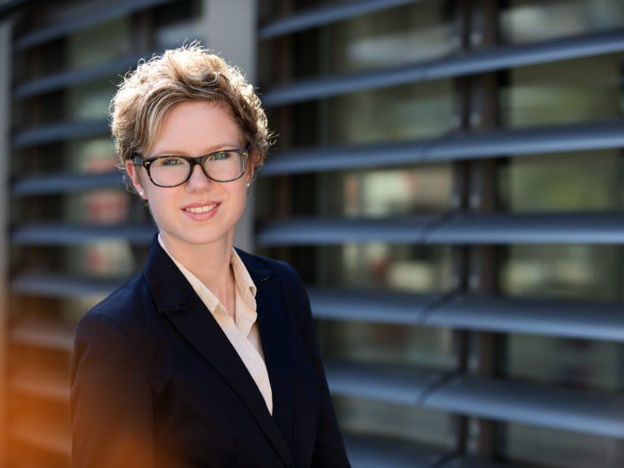 Eine Frau mit Brille und Anzug steht vor einem Gebäude.