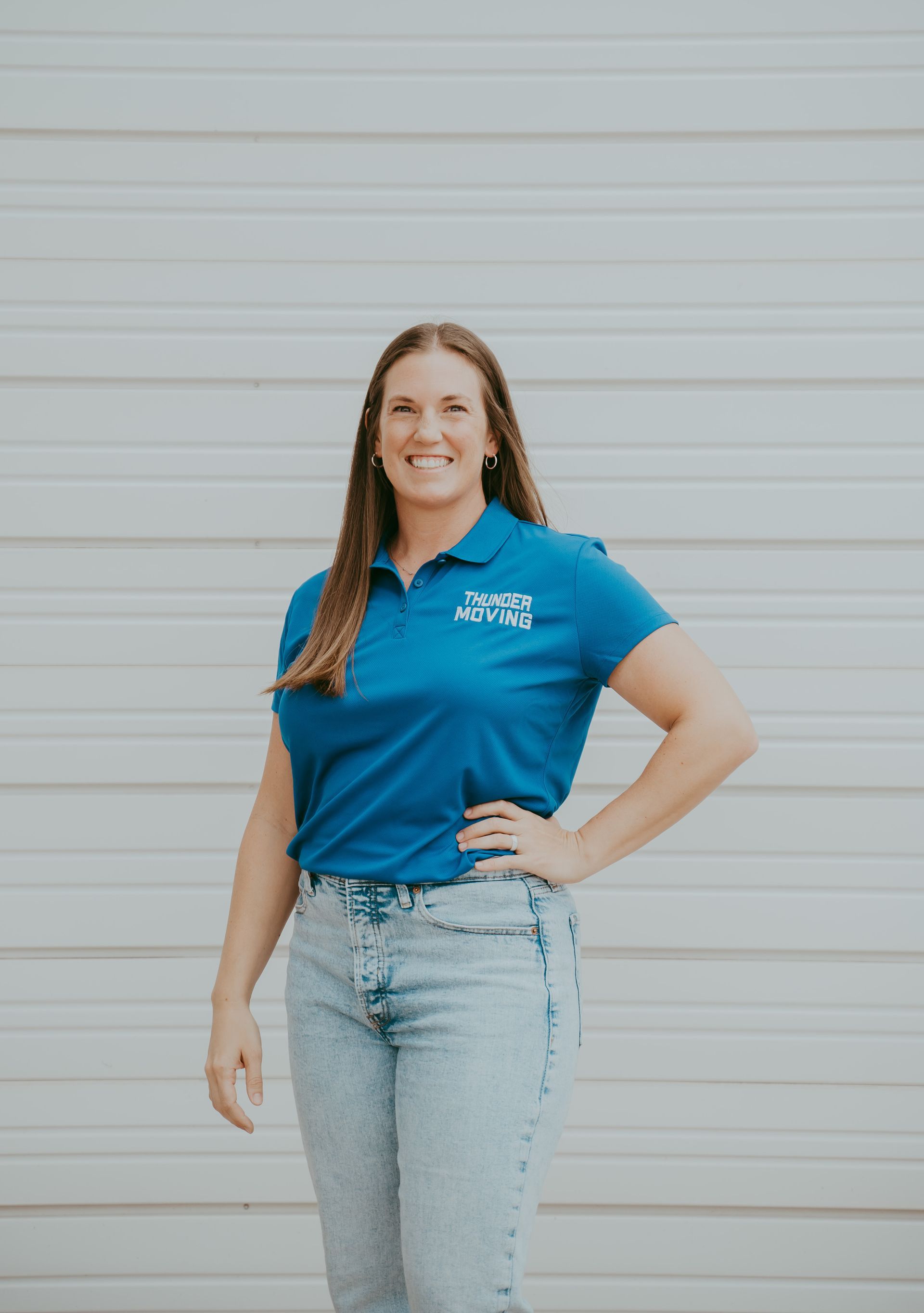 A woman in a blue shirt is smiling and standing in front of a wall.
