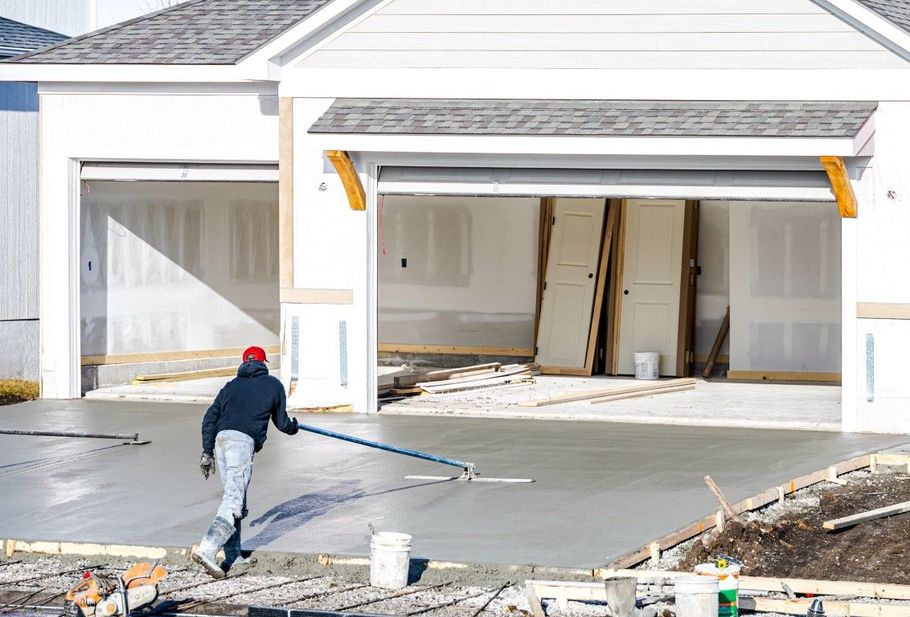 A worker in a hoodie and red hat uses a long-handled tool to smooth wet concrete in front of a house garage.