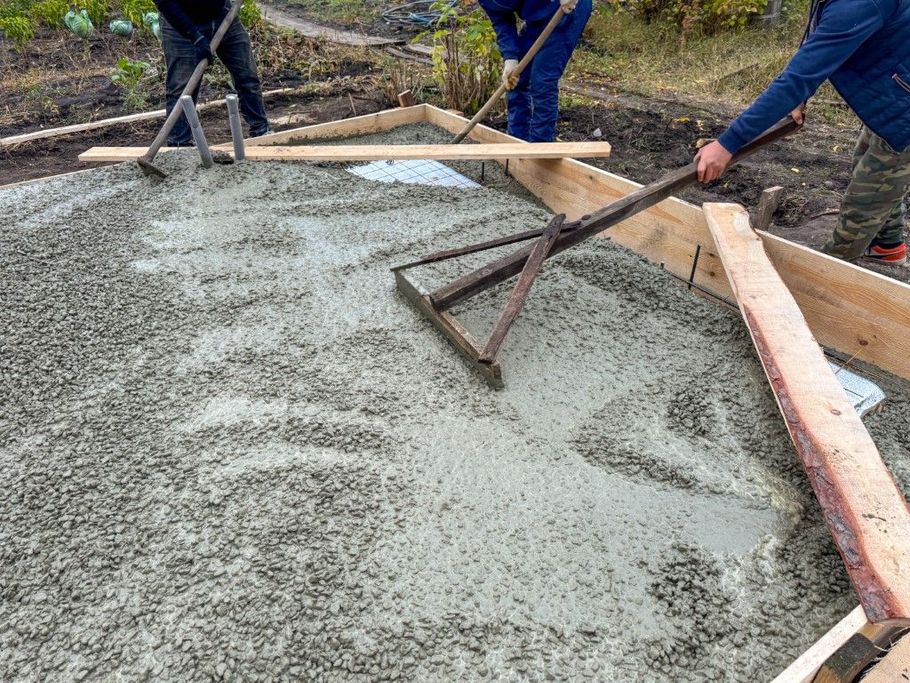 Three people use rakes and boards to level wet concrete within a wooden frame on a construction site.