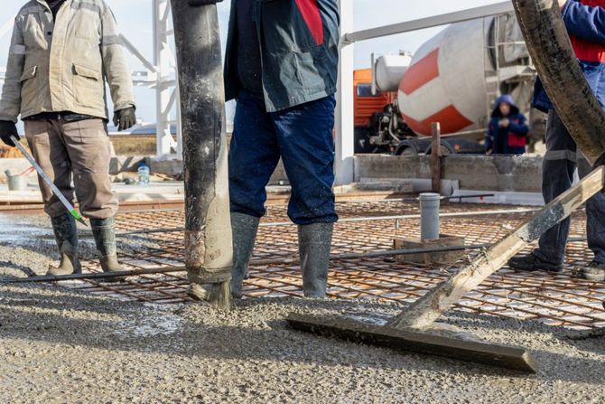 Construction workers pour concrete from a hose onto a reinforced mesh base at a building site.