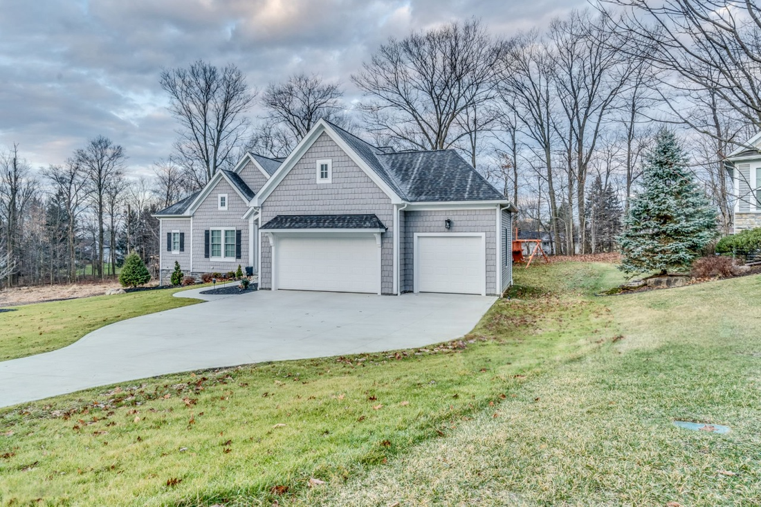 A light gray suburban house with a white three-car garage and concrete driveway surrounded by trees under a cloudy sky.