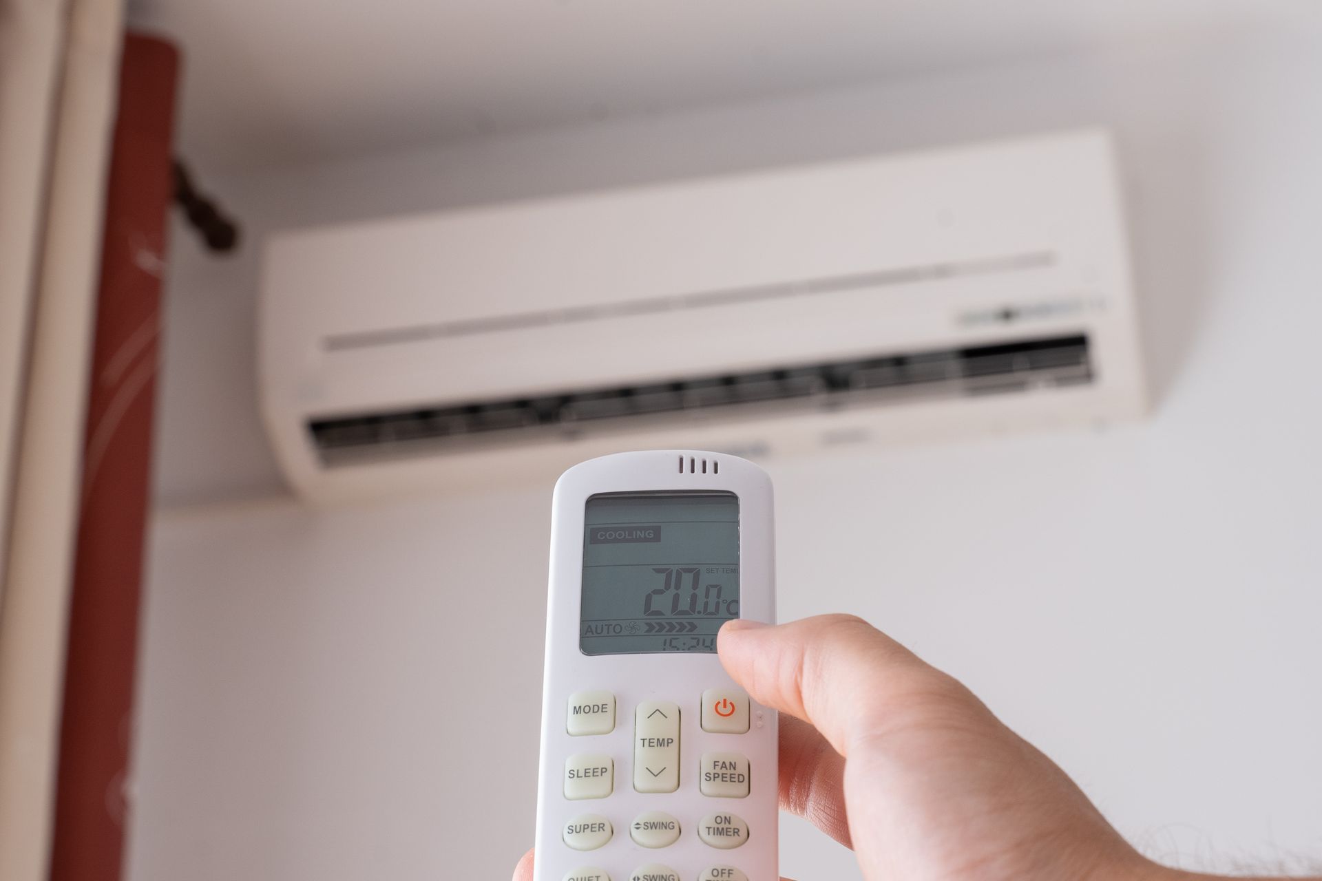 Hand adjusting air conditioner remote, setting temperature to 20°C. Air conditioner unit is mounted on white wall.
