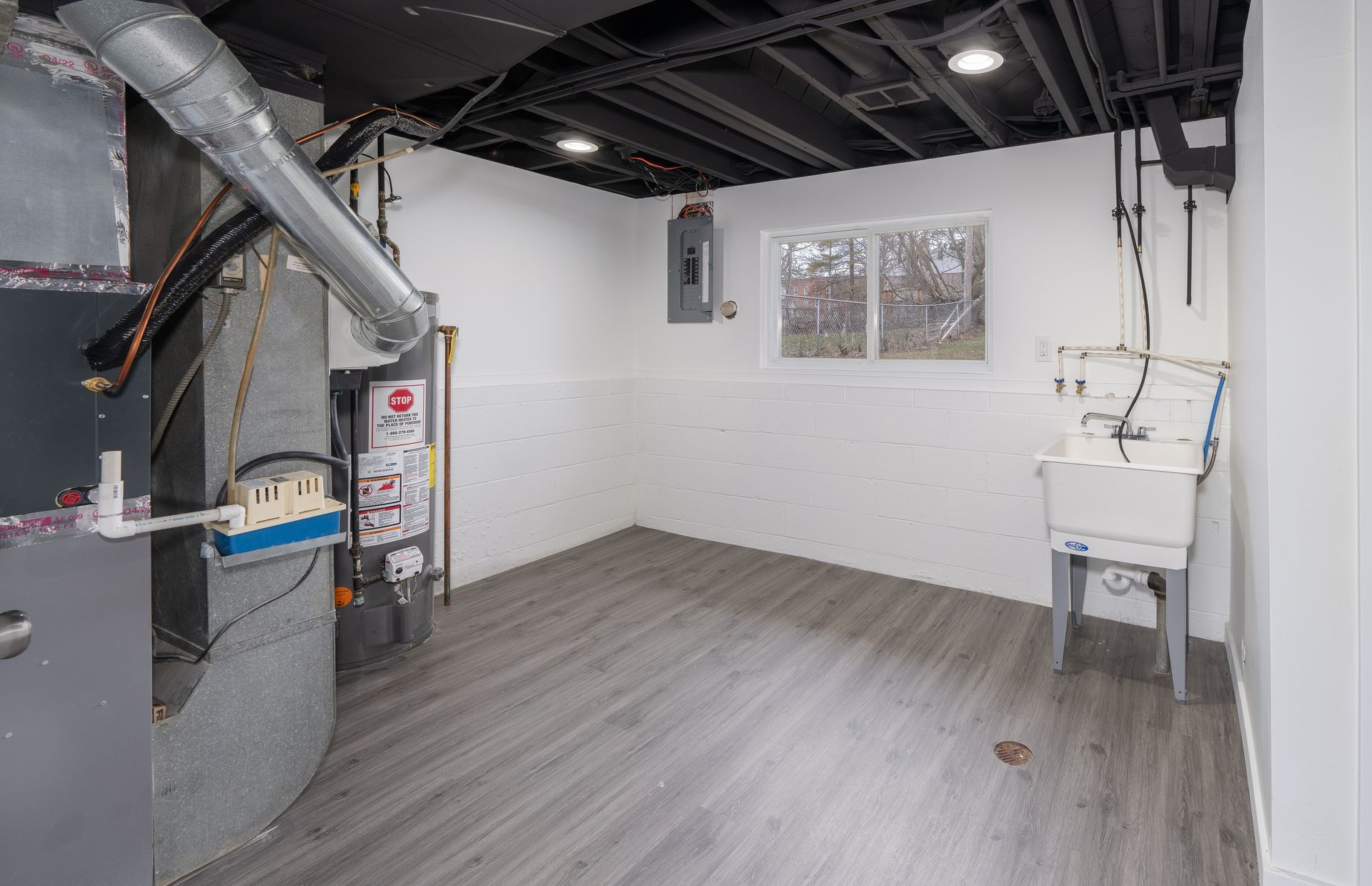 Basement with a furnace, utility sink, electrical panel, window, and gray flooring. White walls and ceiling.