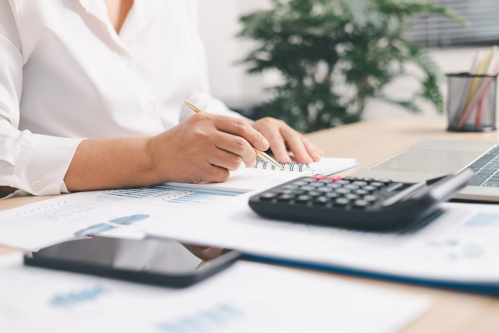 A person in a white shirt sits at a desk with a calculator, smartphone, and financial charts, writing in a notebook.