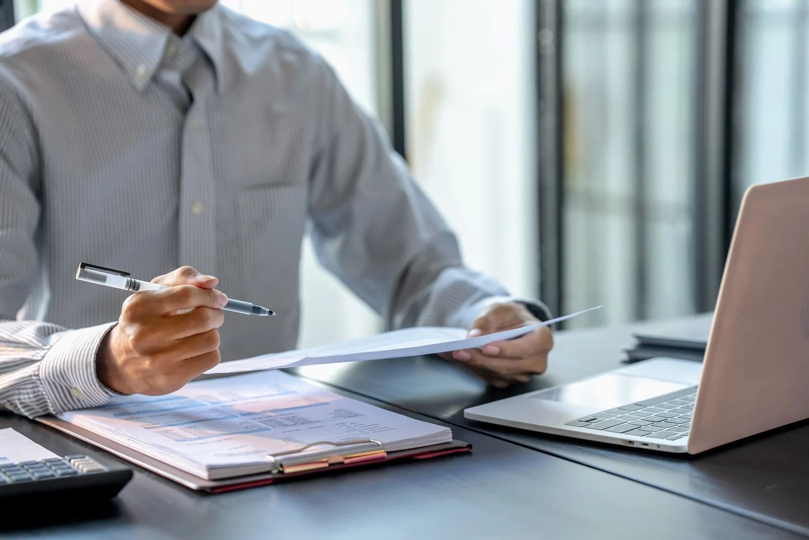 A person in a light shirt sits at a desk with a laptop, holding a pen and reviewing documents on a clipboard.