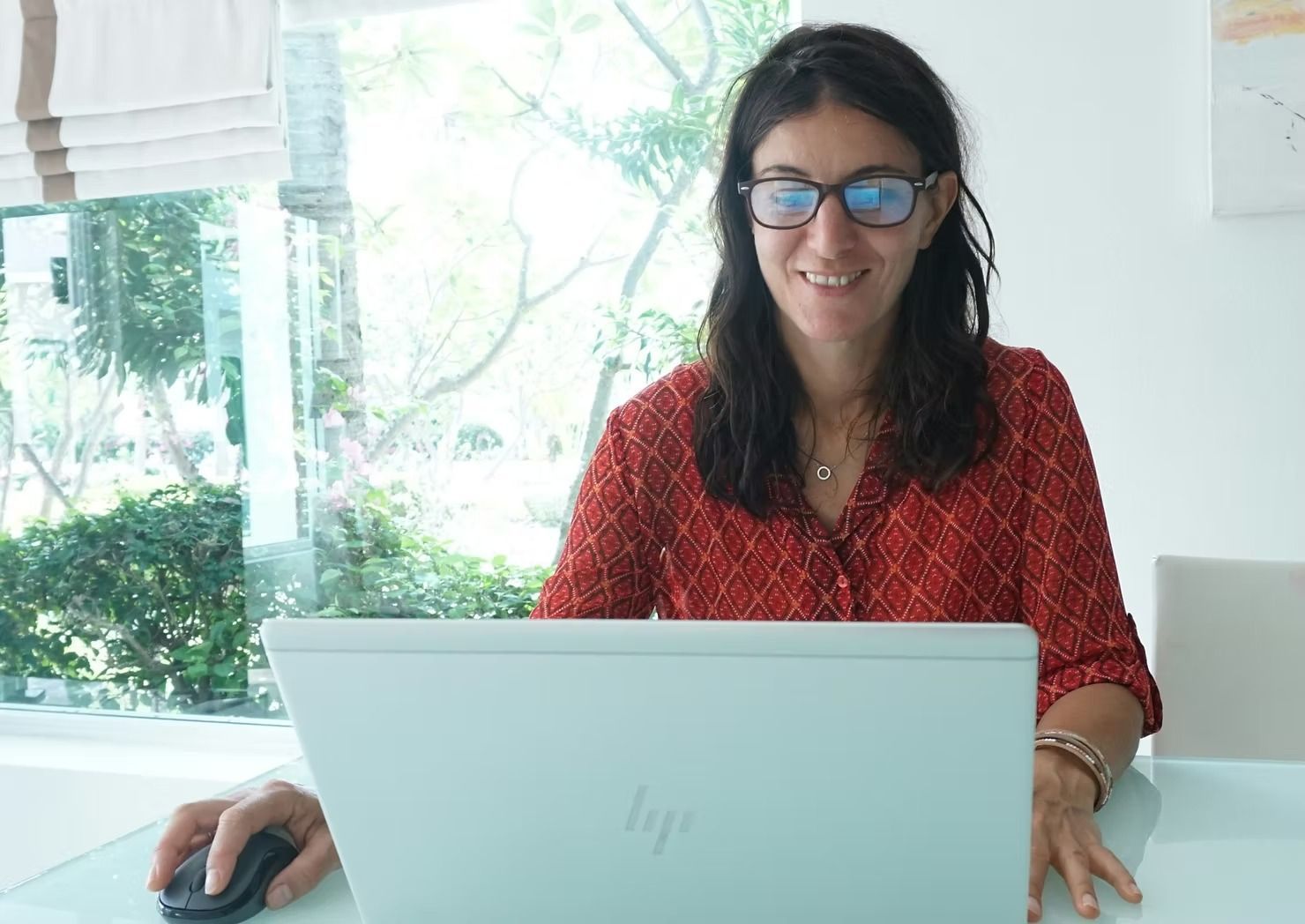 A person wearing glasses and a red patterned shirt smiles while using a laptop and mouse at a bright table.