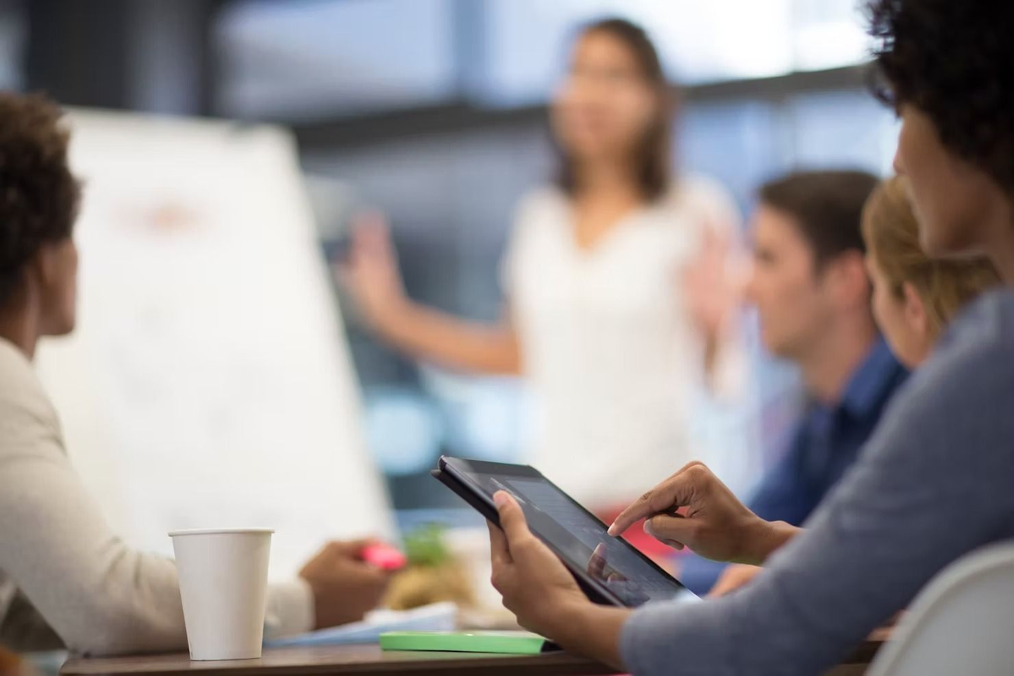 A team meeting where a person uses a tablet in the foreground while another person presents by a whiteboard.