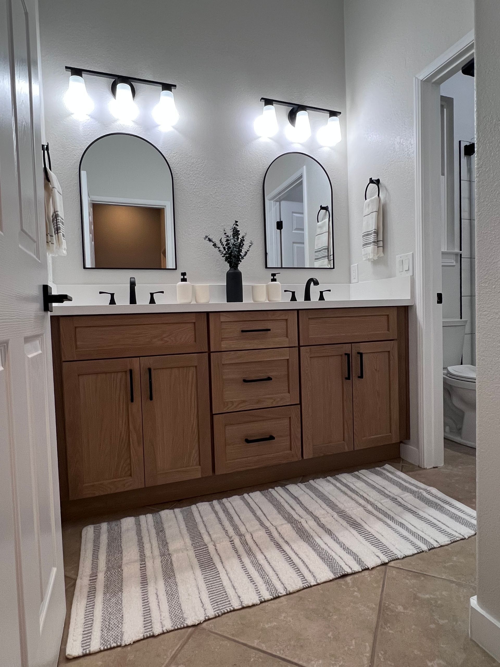 Bathroom with double vanity, arched mirrors, and wood cabinets; neutral tones, rug on floor.
