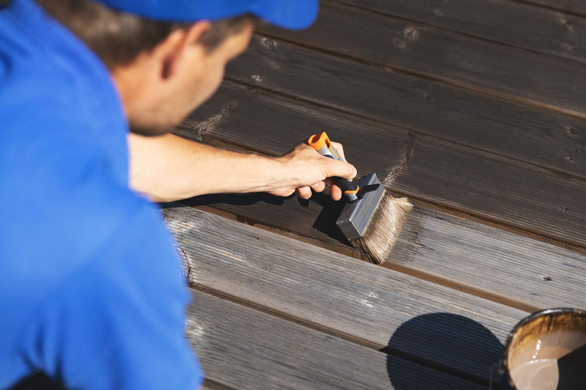 Person in blue painting a dark stain onto wooden deck boards.