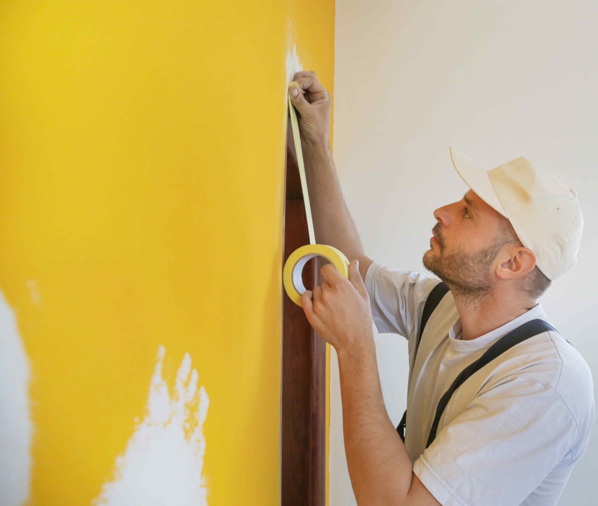 Person taping off wall, preparing to paint. Yellow wall and white wall. Beige hat and shirt.