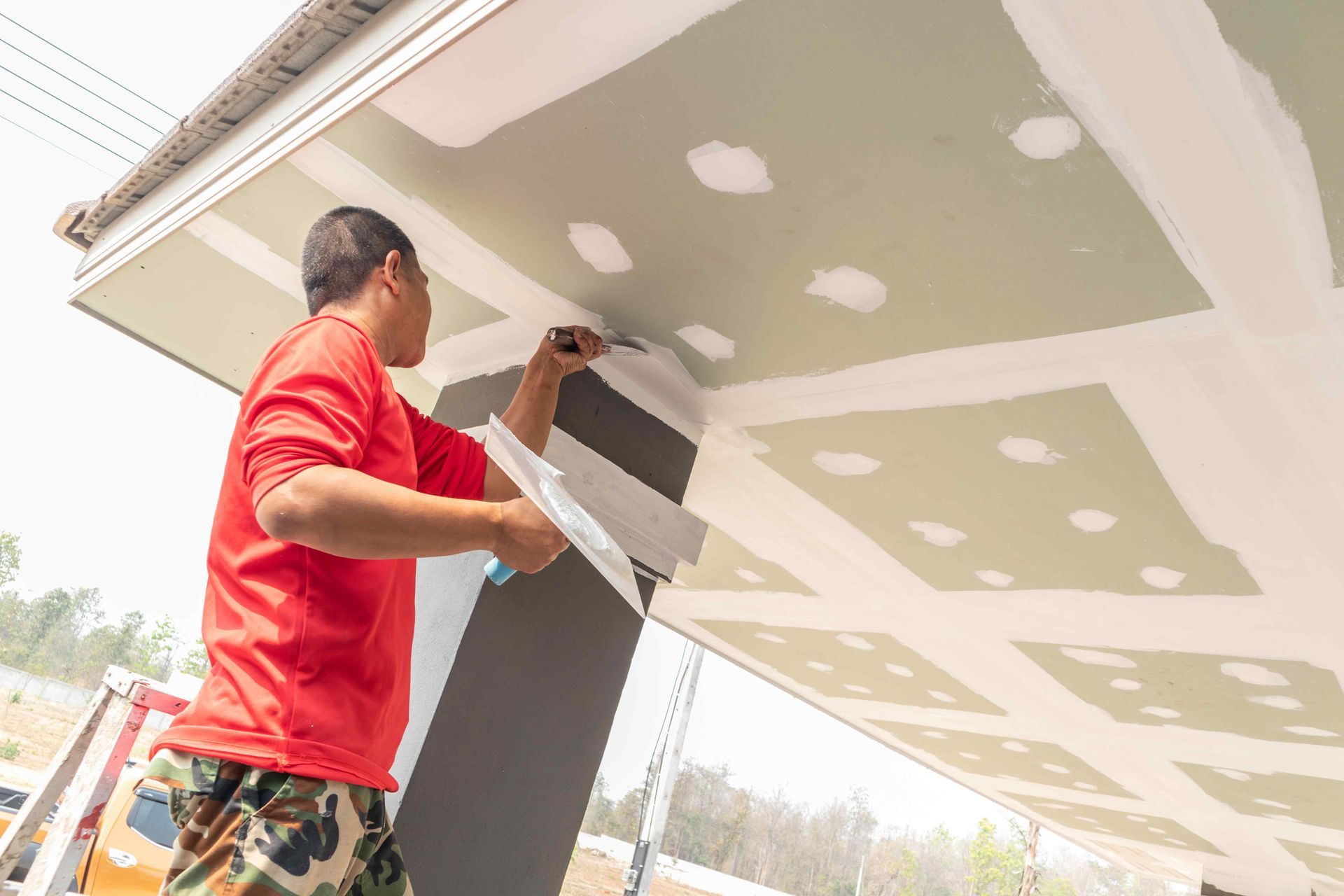 Person in red shirt taping drywall ceiling.