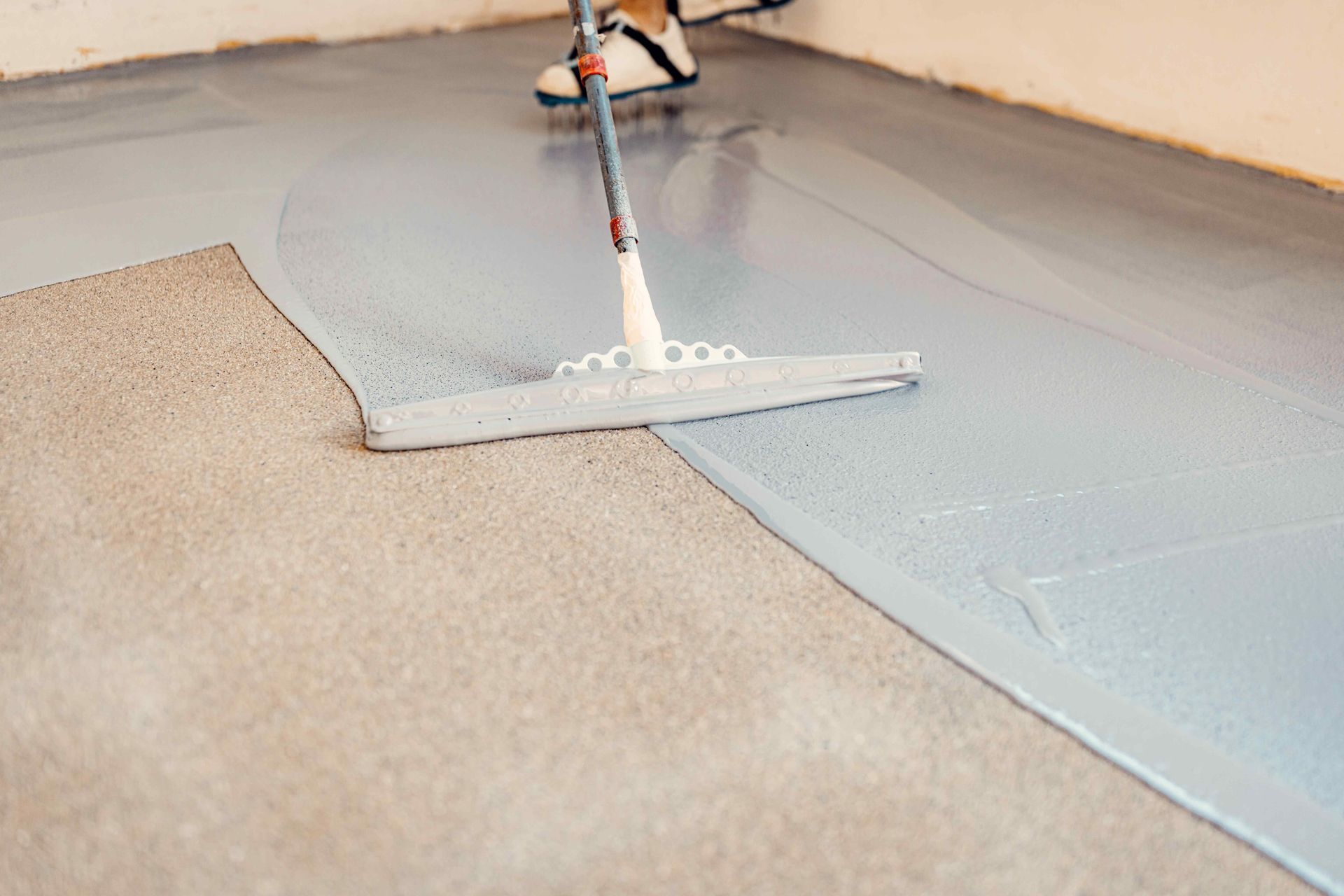 Person using a floor squeegee to apply a grey coating to a concrete floor.