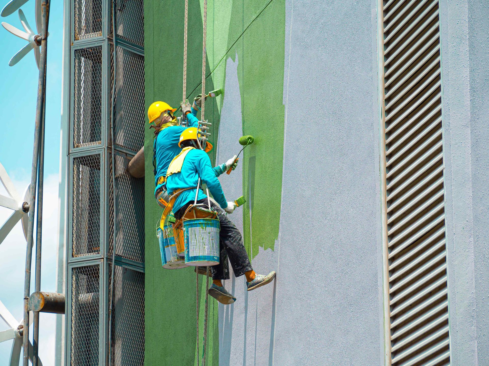 Two painters in safety gear paint a high-rise building wall green, using rollers while suspended.